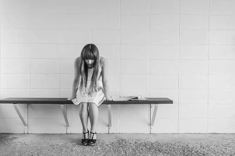 Young woman in a light dress and heels sits on a bench against a tiled wall, head bowed with a folded newspaper beside her