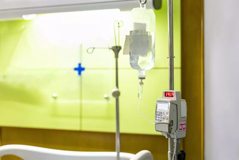 IV drip bag and infusion pump on a pole beside a hospital bed against a green wall with a blue medical cross.