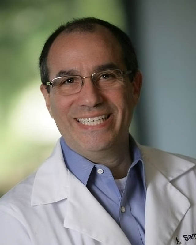 Smiling man wearing glasses and a white lab coat over a blue shirt, blurred green-gray background