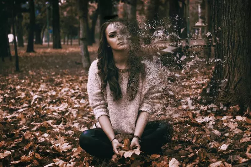 Young woman sitting cross-legged among autumn leaves, her right side dissolving into scattered particle fragments.
