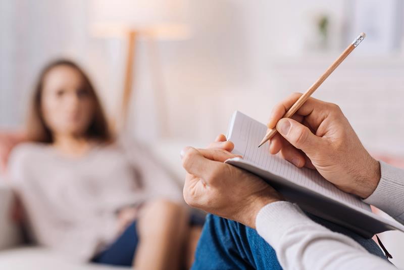 Therapist's hands writing in a notepad with pencil while a patient reclines blurred in the background.